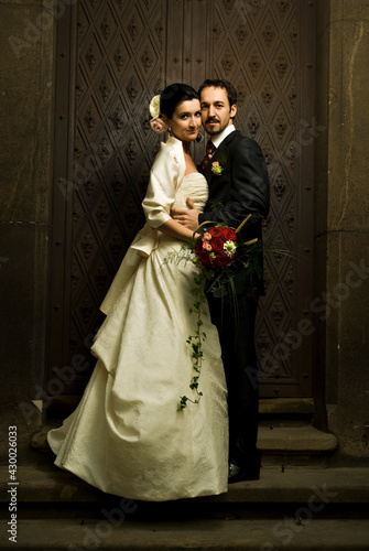 Gorgeous wedding couple (bride and groom) standing on staircase in front of historical church doorway and looking at camera.
