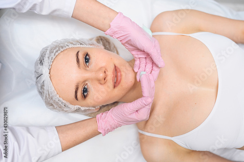 Beautiful happy woman receiving spa treatment. Cosmetologist in beauty salon with a pink glove applies a moisturizing cream mask. Facial beauty. Perfect fresh clean skin. Youth and skin care concept 