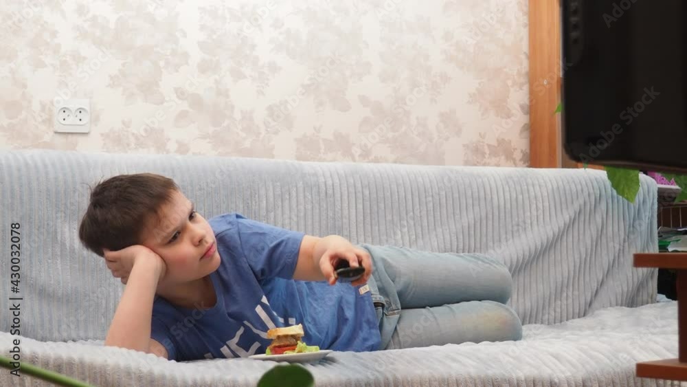 a teenage boy at home lies on the couch and watches TV, cartoons or movies, switches channels, programs. Eating a hamburger. Free time. Leisure. modern youth a teenage boy at home lies on the couch and watches TV, cartoons or movies, switches channels, programs. Eating a hamburger. Free time. Leisure. modern youth