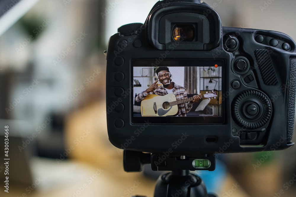 Cheerful young man with dark skin recording video on camera while playing guitar. Happy male blogger creating music content for his social networks. Cheerful young man with dark skin recording video on camera while playing guitar. Happy male blogger creating music content for his social networks.