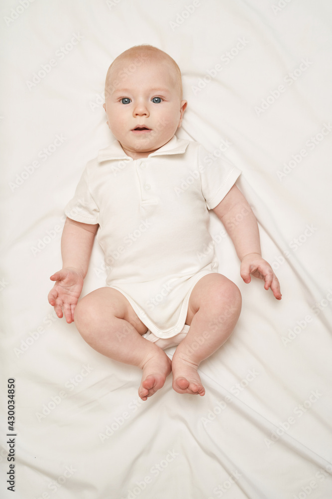Cute small boy lying at bed. Childhood concept. Light background. Serious child. Copyspace. Stay home. Onesie mockup. White clothes. Full body Cute small boy lying at bed. Childhood concept. Light background. Serious child. Copyspace. Stay home. Onesie mockup. White clothes. Full body