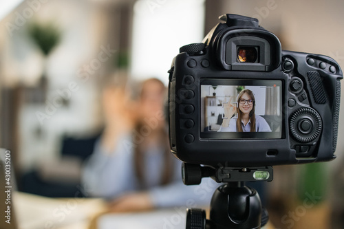 Smiling female influencer using modern camera for leading live stream while sitting at home. Focus on screen of video camera. Concept of people and technology.