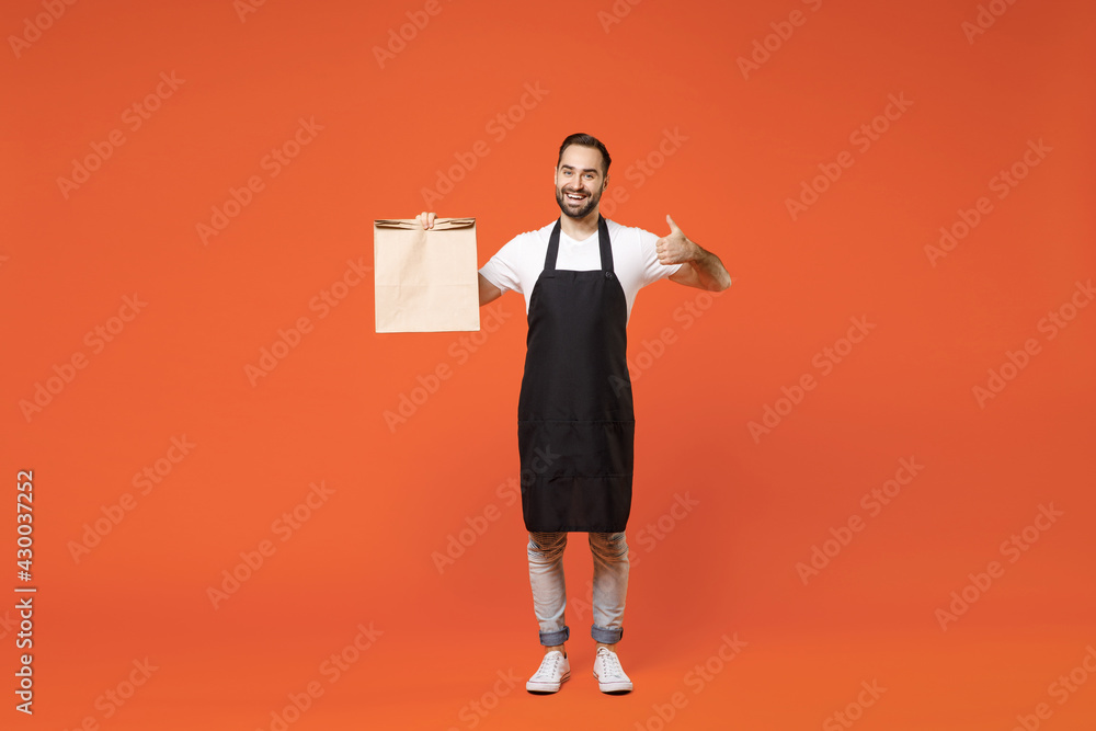 Full length young man barista bartender barman employee in apron white tshirt work coffee shop hold blank craft paper takeaway bag show thumb up gesture isolated on orange background Business startup Full length young man barista bartender barman employee in apron white tshirt work coffee shop hold blank craft paper takeaway bag show thumb up gesture isolated on orange background Business startup