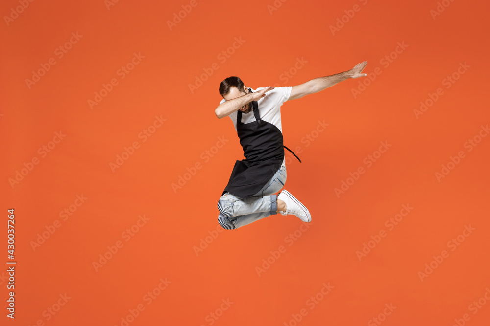 Full length young man barista bartender barman employee in apron white t-shirt work in coffee shop do dab hip hop dance hands gesture hiding face isolated on orange background. Small business startup.