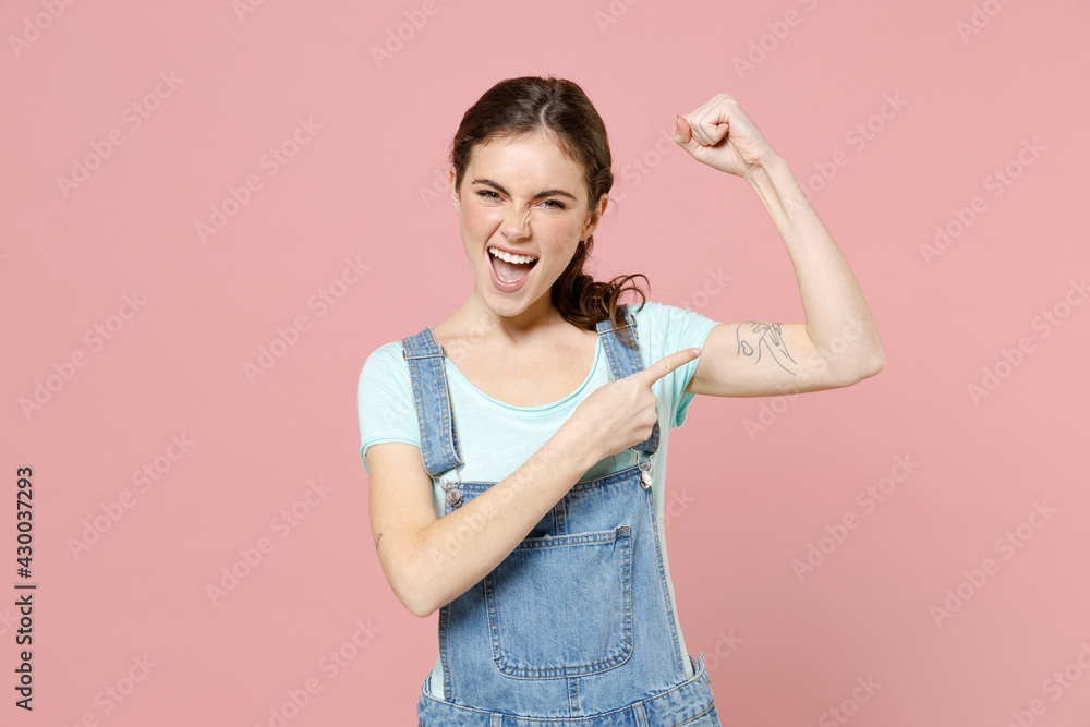 Young strong sporty fitness woman wear trendy stylish denim clothes blue t-shirt point finger on biceps muscles on hand demonstrating strength power isolated on pastel pink background studio portrait Young strong sporty fitness woman wear trendy stylish denim clothes blue t-shirt point finger on biceps muscles on hand demonstrating strength power isolated on pastel pink background studio portrait