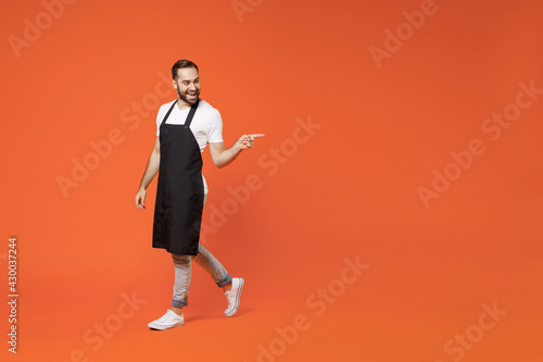 Full length young man barista bartender barman employee in black apron white tshirt work in coffee shop point finger aside area workspace isolated on orange background. Small business startup concept.