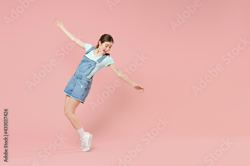 Full length young smiling joyful caucasian woman 20s in trendy denim clothes blue t-shirt stand on toes leaning back dancing with outstretched hands isolated on pastel pink background studio portrait.