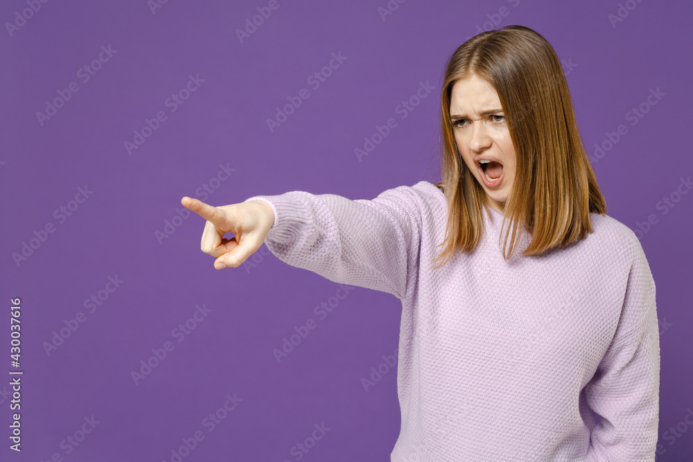 © ViDi Studio - Young angry strict indignant student woman in purple knitted sweater point index finger aside scream shout command do it isolated on violet color background studio portrait People lifestyle concept.