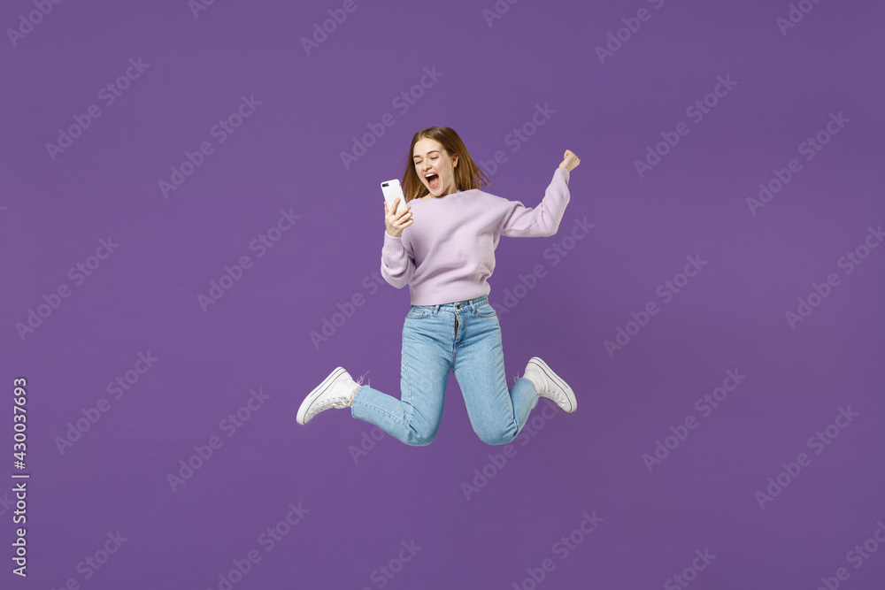 Full length young excited overjoyed student caucasian happy woman 20s wear purple knitted sweater jump high hold mobile cell phone isolated on violet color background studio People lifestyle concept.