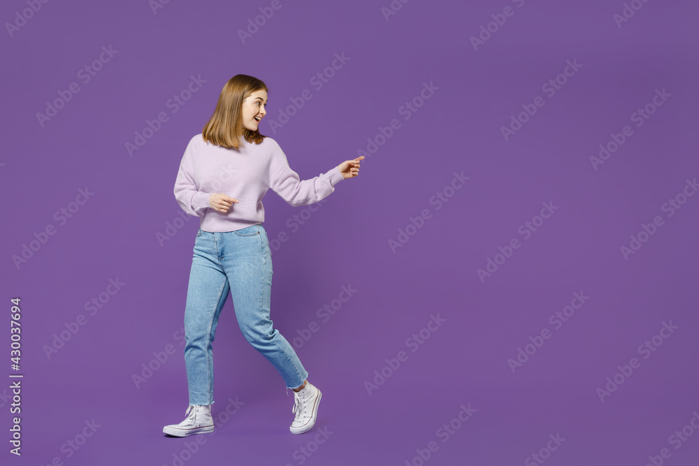 Full length young surprised happy student woman 20s wear purple knitted sweater walk go point index finger aside on copy space isolated on violet background studio portrait People lifestyle concept