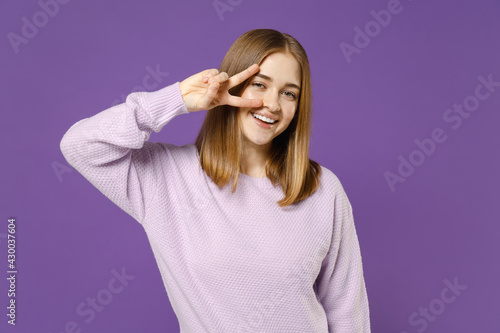Young smiling positive happy cheerful student woman 20s wear purple knitted sweater cover eye with victory v-sign gesture isolated on violet color background studio portrait People lifestyle concept.