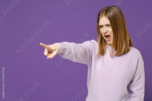 Young angry strict indignant student woman in purple knitted sweater point index finger aside scream shout command do it isolated on violet color background studio portrait People lifestyle concept. photo