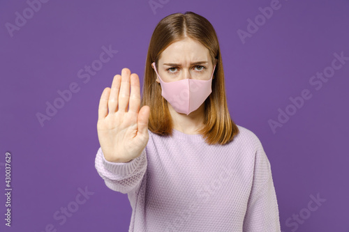 Young sad woman 20s in purple knitted sweater fabric face mask safe from coronavirus virus covid-19 during pandemic quarantine do stop palm gesture isolated on violet color background studio portrait.