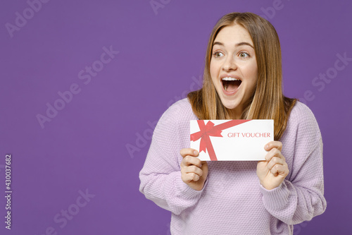 Young fun surprised excited happy student woman 20s wearing purple knitted sweater hold gift voucher flyer mock up look aside isolated on violet background studio portrait. People lifestyle concept