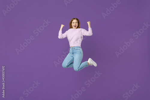 Full length young excited overjoyed happy woman 20s wearing purple knitted sweater jump high do winner gesture clench fist isolated on violet color background studio portrait People lifestyle concept.
