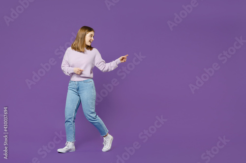 Full length young surprised happy student woman 20s wear purple knitted sweater walk go point index finger aside on copy space isolated on violet background studio portrait People lifestyle concept