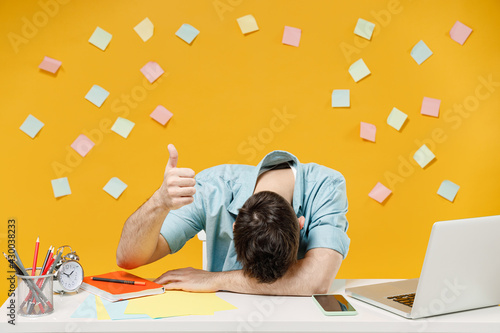 Young tired exhausted sarcastic sleepy employee business man wear shirt sit work at white office desk with laptop sleep put head on table show thumb up isolated on yellow background studio portrait photo