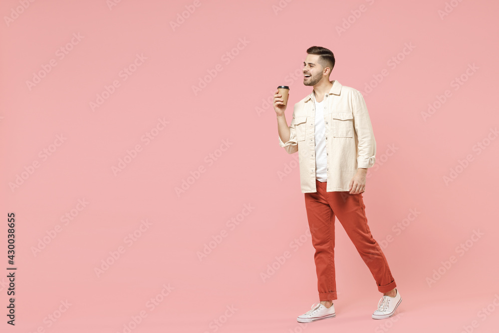 Full length young smiling happy fun trendy fashionable friendly caucasian man 20s wearing jacket white t-shirt drink hot coffee hold paper cup walk isolated on pastel pink color background studio. Full length young smiling happy fun trendy fashionable friendly caucasian man 20s wearing jacket white t-shirt drink hot coffee hold paper cup walk isolated on pastel pink color background studio.
