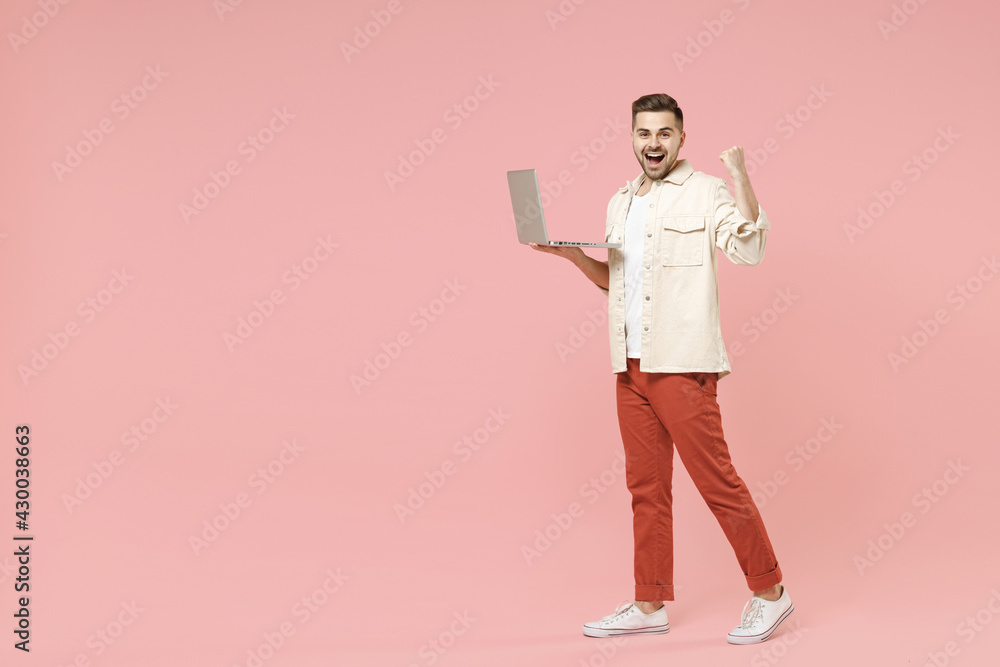 Full length young overjoyed happy fun fashionable friendly man in jacket white t-shirt using laptop pc computer do winner gesture clench fist celebrating isolated on pastel pink background studio. Full length young overjoyed happy fun fashionable friendly man in jacket white t-shirt using laptop pc computer do winner gesture clench fist celebrating isolated on pastel pink background studio.