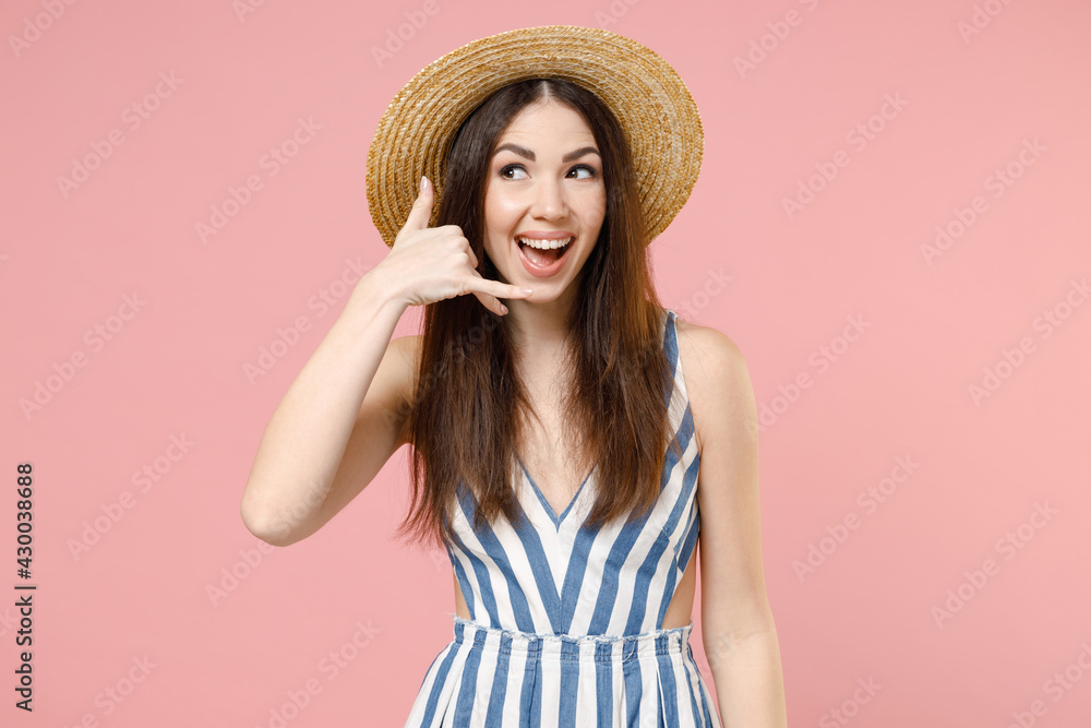 Young excited cheerful caucasian woman in summer clothes striped dress straw hat doing phone gesture like says call me back isolated on pastel pink background studio portrait People lifestyle concept