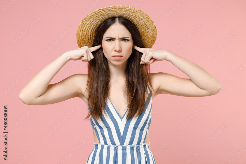 Young irritated sad angry nervous stressed caucasian woman in summer clothes striped dress straw hat cover ears with fingers hands isolated on pastel pink background studio. People lifestyle concept. Young irritated sad angry nervous stressed caucasian woman in summer clothes striped dress straw hat cover ears with fingers hands isolated on pastel pink background studio. People lifestyle concept.
