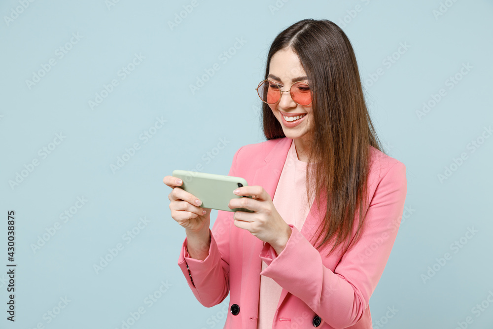 Young gambling woman in pastel pink clothes glasses use play racing mobile cell phone hold gadget smartphone for pc video games isolated on blue background studio People lifestyle technology concept. Young gambling woman in pastel pink clothes glasses use play racing mobile cell phone hold gadget smartphone for pc video games isolated on blue background studio People lifestyle technology concept.