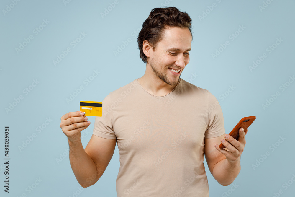 Young rich happy smiling excited caucasian man 20s wear casual basic beige t-shirt hold mobile cell phone credit bank card isolated on pastel blue background studio portrait. People lifestyle concept. Young rich happy smiling excited caucasian man 20s wear casual basic beige t-shirt hold mobile cell phone credit bank card isolated on pastel blue background studio portrait. People lifestyle concept.