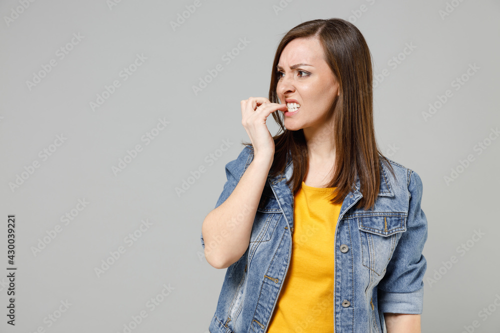 Fototapeta premium Young troubled pensive puzzled indignant caucasian woman 20s wear casual denim jacket yellow t-shirt look aside biting nails isolated on grey color background studio portrait People lifestyle concept.