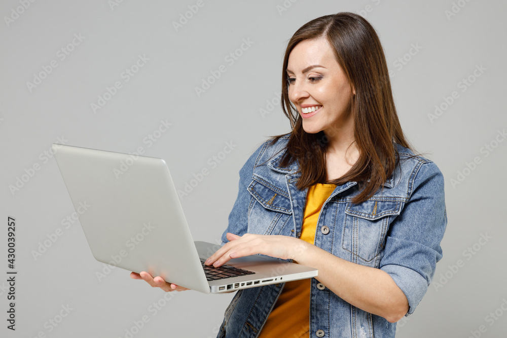 Young smiling happy caucasian woman in casual trendy denim jacket yellow t-shirt using laptop pc computer browsing chat online isolated on grey background studio portrait. People lifestyle concept Young smiling happy caucasian woman in casual trendy denim jacket yellow t-shirt using laptop pc computer browsing chat online isolated on grey background studio portrait. People lifestyle concept