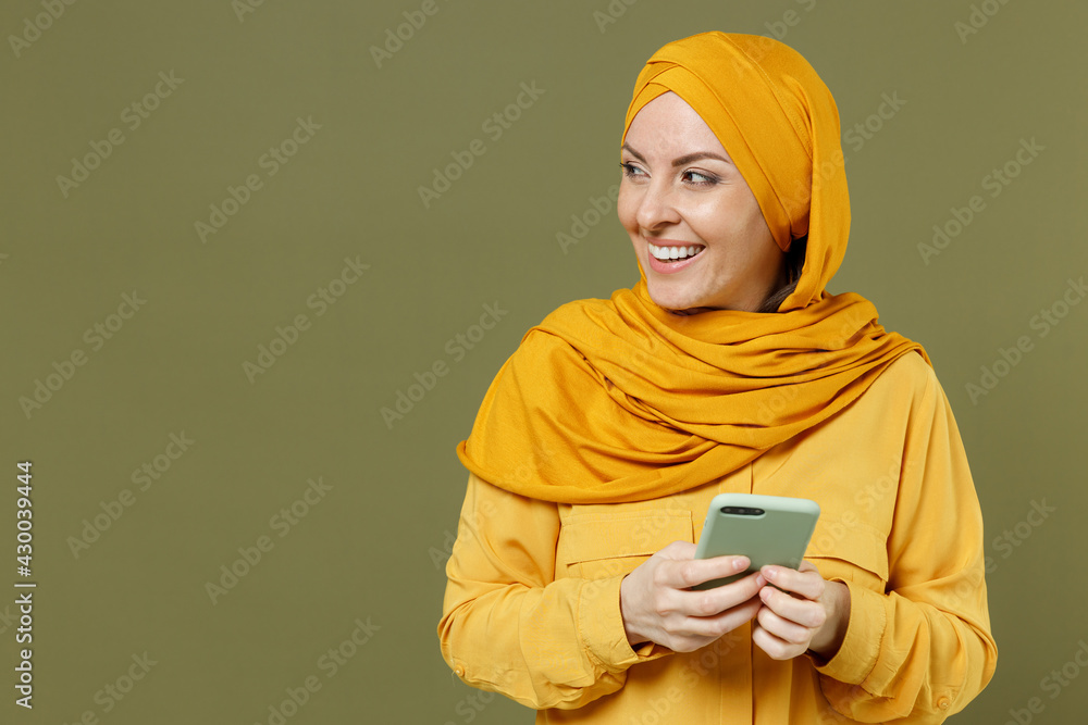 Smiling excited young arabian asian muslim woman in abaya hijab yellow clothes using mobile cell phone look aside isolated on olive green background studio portrait People uae islam religious concept Smiling excited young arabian asian muslim woman in abaya hijab yellow clothes using mobile cell phone look aside isolated on olive green background studio portrait People uae islam religious concept