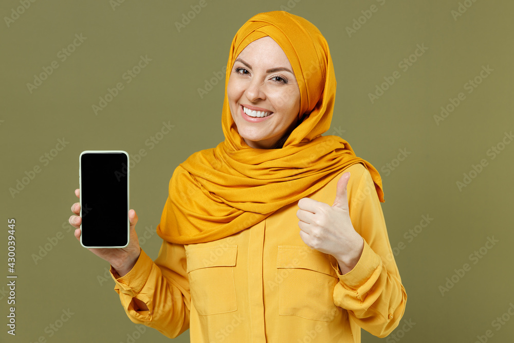 Young fun arabian asian muslim woman in abaya hijab yellow clothes hold mobile cell phone with blank workspace area show thumb up isolated on olive green background People uae islam religious concept Young fun arabian asian muslim woman in abaya hijab yellow clothes hold mobile cell phone with blank workspace area show thumb up isolated on olive green background People uae islam religious concept