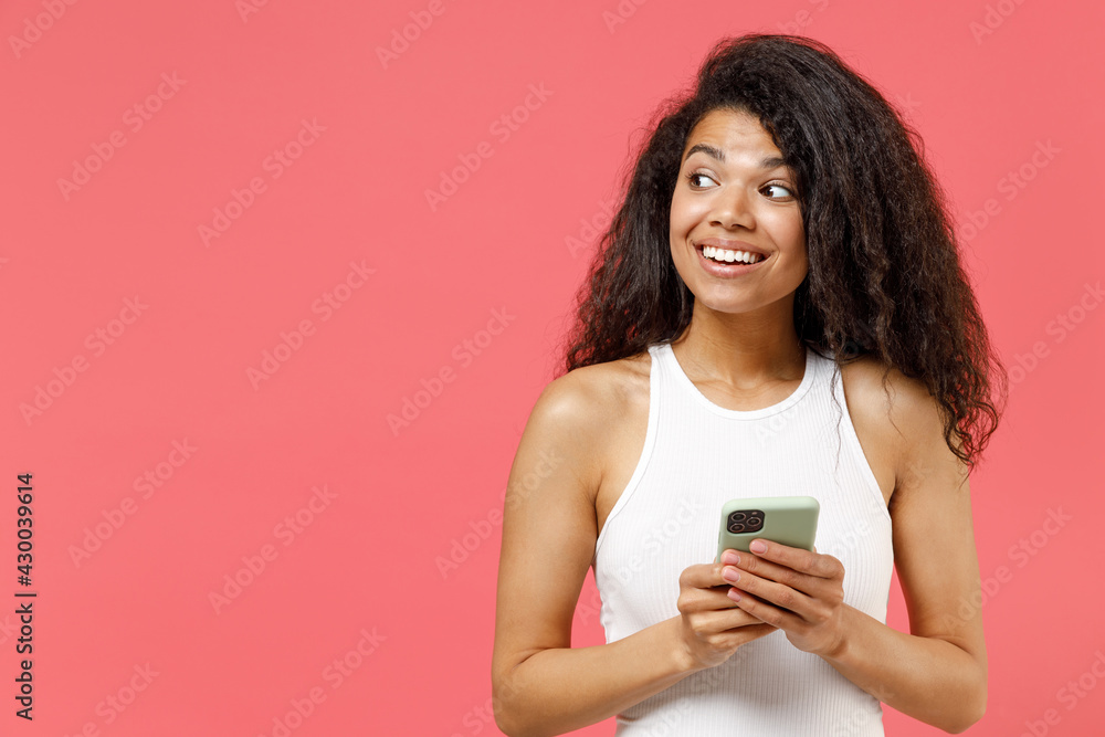 Young smiling happy dreamful thoughtful wistful pensive african american woman 20s wear casual white tank shirt hold in hand using mobile cell phone chat online look aside isolated on pink background Young smiling happy dreamful thoughtful wistful pensive african american woman 20s wear casual white tank shirt hold in hand using mobile cell phone chat online look aside isolated on pink background