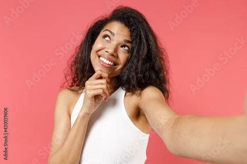 Close up young dreamful pensive wistful thoughtful african american woman wear white tank shirt doing selfie shot on mobile phone prop up chin look aside isolated on pink background studio portrait