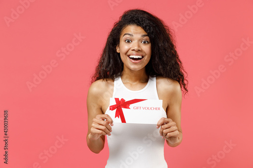 Young fun smiling excited happy satisfied positive african american woman 20s wearing casual white tank shirt hold in hand gift voucher flyer mock up isolated on pink color background studio portrait.