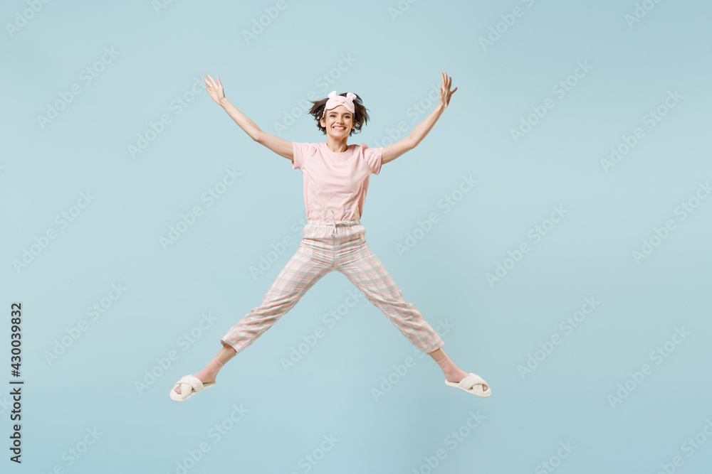 Full length young overjoyed woman in pajamas jam sleep eye mask rest relax at home jump high do winner gesture clench fist isolated on pastel blue background studio. Good mood night bedtime concept.