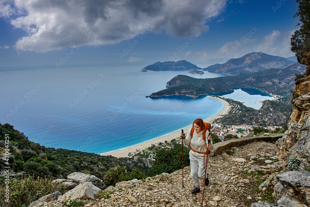 Blue lagoon in Oludeniz near Fethiye in Turkey, young woman climbs Lycian path using trekking poles.