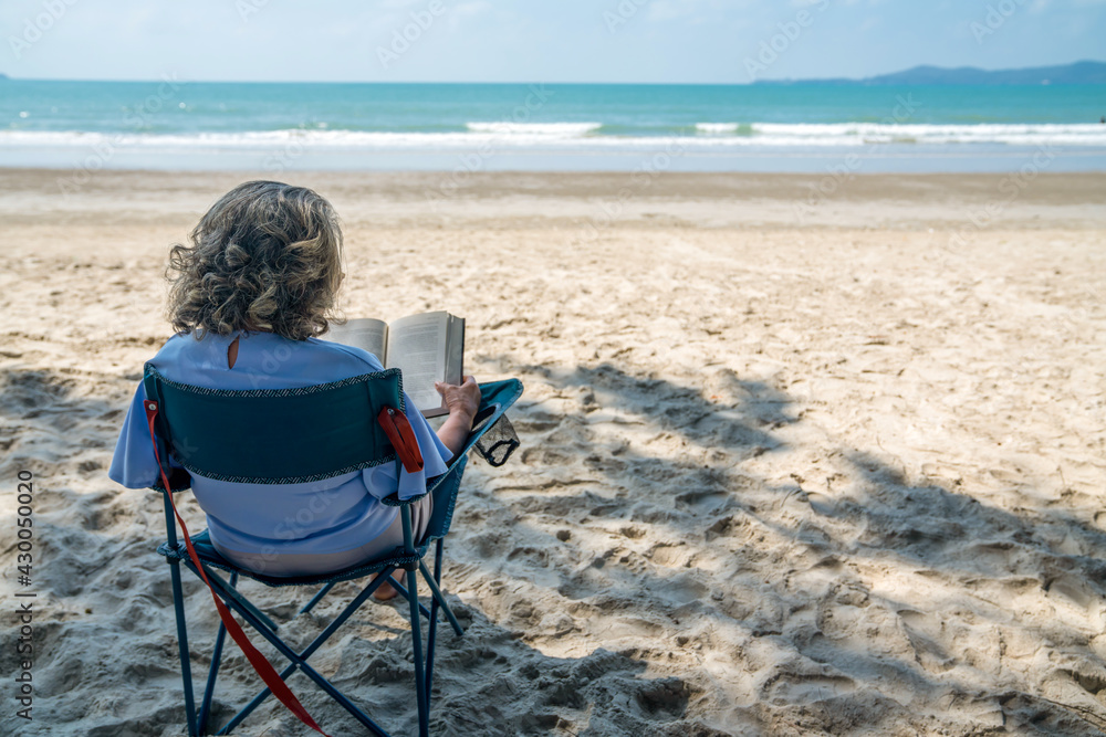 Happy Asian senior woman sitting on outdoor chair on the beach with reading a book. Healthy retirement elderly female resting by the sea relax and enjoy outdoor lifestyle activity in summer vacation
