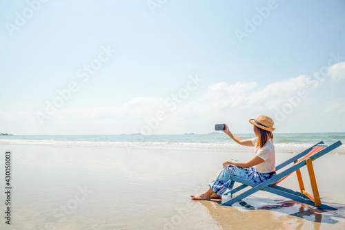 Asian woman resting on sunbed on the beach. Happy girl sit on beach chair using smartphone selfie or video call with friend. Female relax and enjoy beach outdoor lifestyle activity on summer vacation © CandyRetriever 