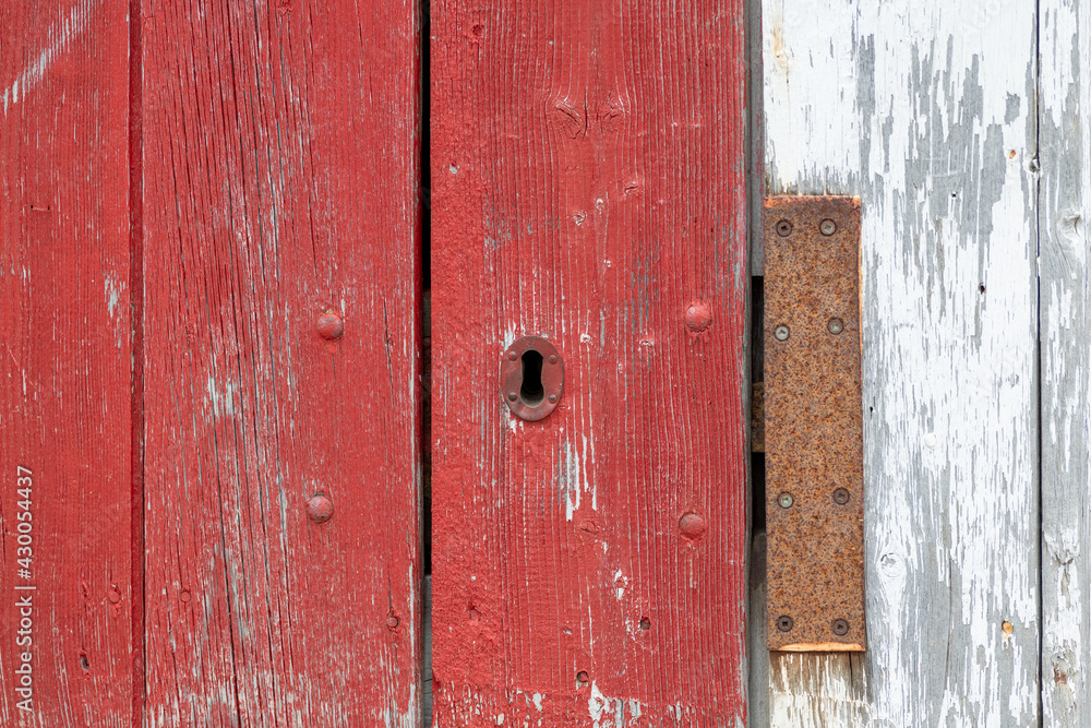 A closeup of a bright red vintage metal keyholder in a textured red wooden door. The exterior of the old woodshed has worn and wear patterns with some scuff marks.The door is on a white wood building.