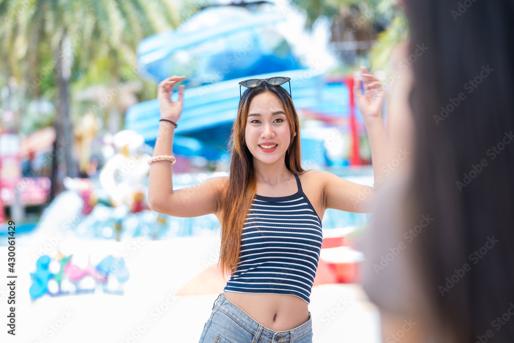 Young asian joyful woman in summer fashion dress standing and posing for  friend take a shoot photo on smartphone in amusement park . Concept happy  and lovely life of teenager. Stock-Foto