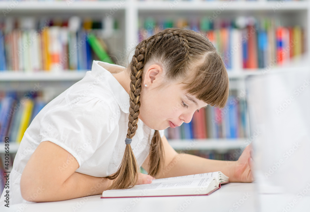 Smart young girl with syndrome down reads a book at library. Education for disabled children concept