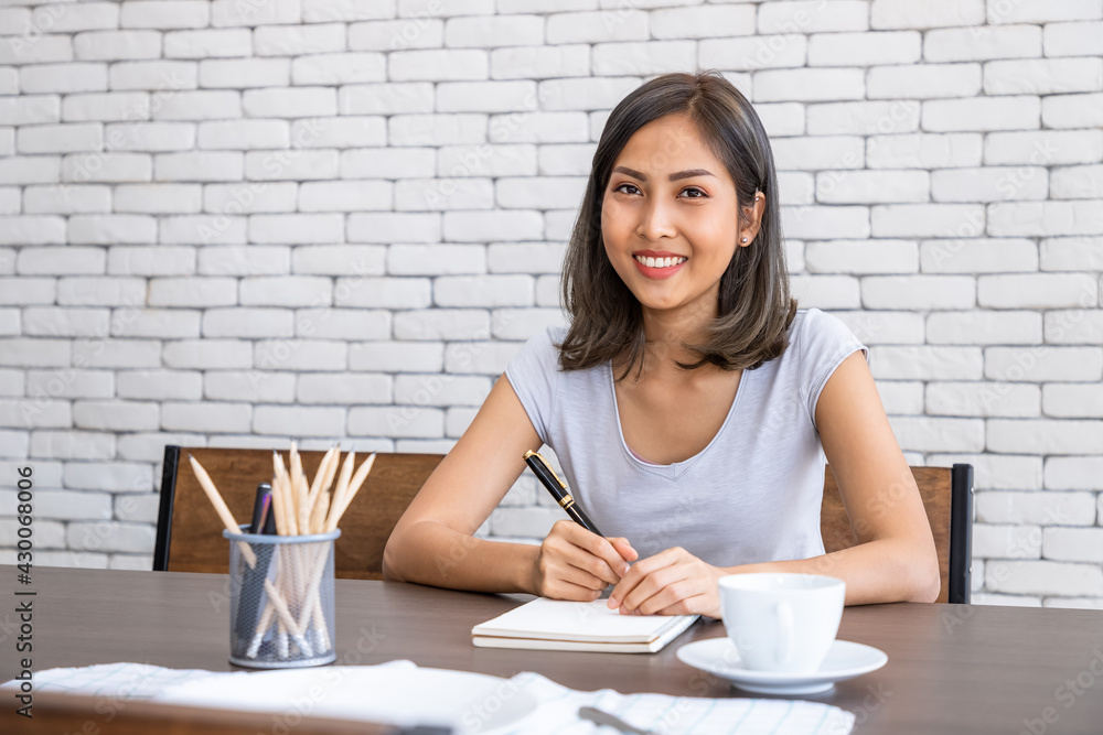 Young attractive Asian woman smiling thinking planning writing memo in notebook working from home, looking at camera Young attractive Asian woman smiling thinking planning writing memo in notebook working from home, looking at camera