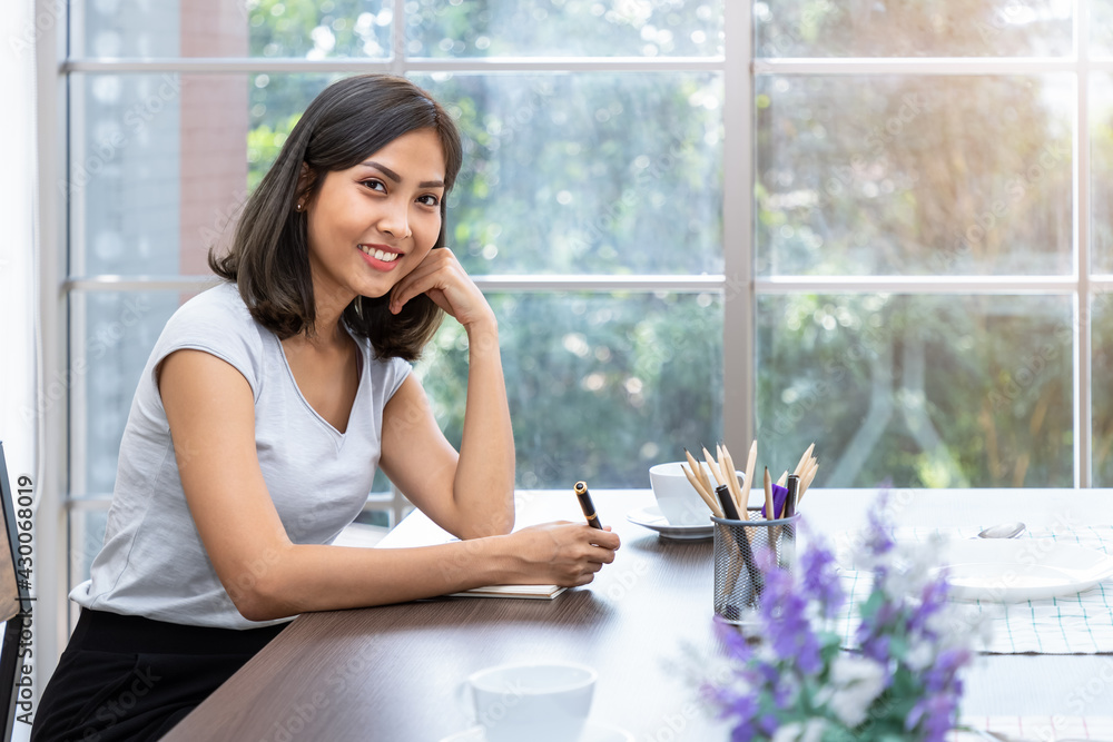 Obraz premium Young attractive Asian woman smiling thinking planning writing memo in notebook working from home, looking at camera