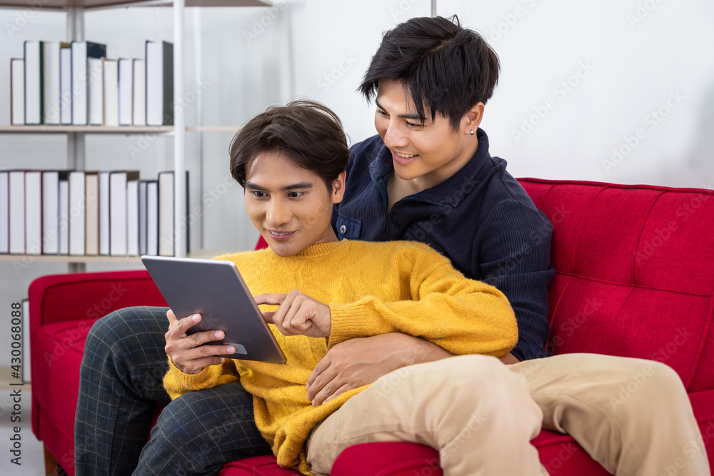 Asian gay couple sitting on sofa using tablet browsing Internet for online shopping, hand around his partner, hugging or embracing. Looking at screen.