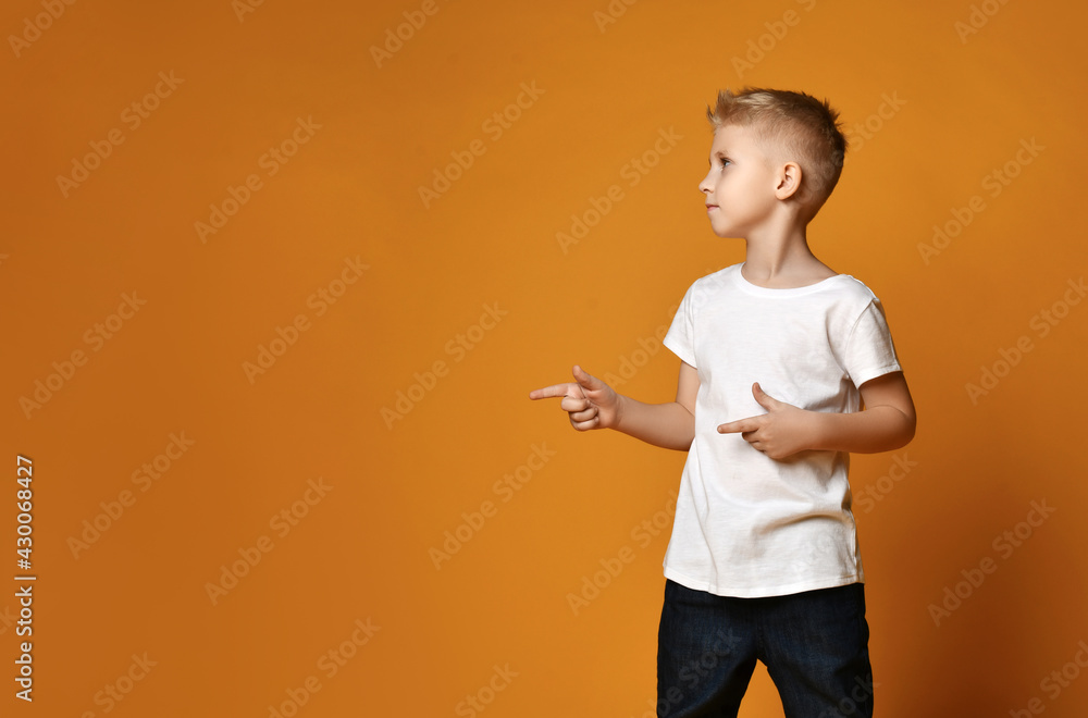 Blond kid boy, teenager, regular guy in white t-shirt stands looking and pointing forefingers aside at copy space over yellow background