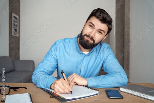 Millennial man student studying online writing in notebook looking at camera