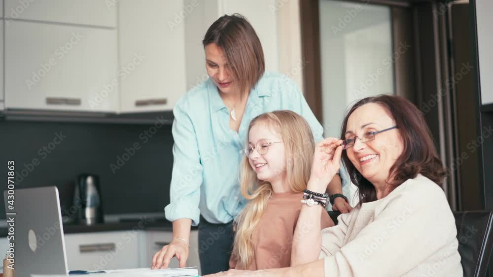 A grandmother in eyeglasses and a mother helping daughter with her homework. Beautiful 10-year-old girl doing homework on a laptop with the support of her mom and grandma. A grandmother in eyeglasses and a mother helping daughter with her homework. Beautiful 10-year-old girl doing homework on a laptop with the support of her mom and grandma.