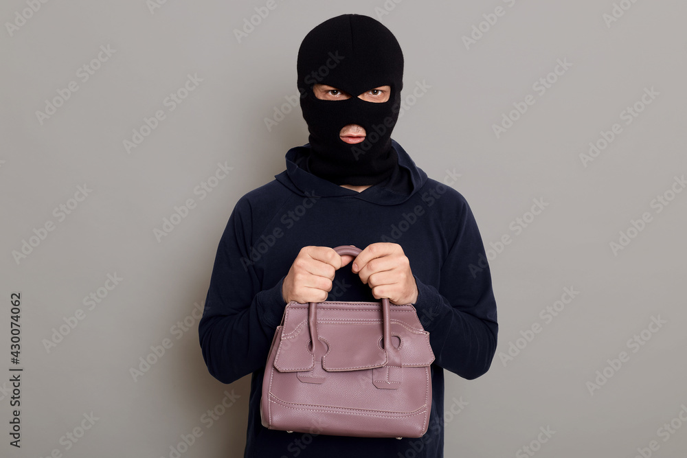 A young guy thief is dressed in a black hoodie and robbery mask, holds a woman's bag with both hands, stole other people's things, looks at the camera, isolated on gray background. A young guy thief is dressed in a black hoodie and robbery mask, holds a woman's bag with both hands, stole other people's things, looks at the camera, isolated on gray background.