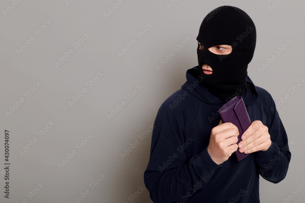 Frightened guy thief dressed in a black hoodie, stole a wallet and runs away looking back, afraid to be caught, copy space, isolated on gray background. Frightened guy thief dressed in a black hoodie, stole a wallet and runs away looking back, afraid to be caught, copy space, isolated on gray background.