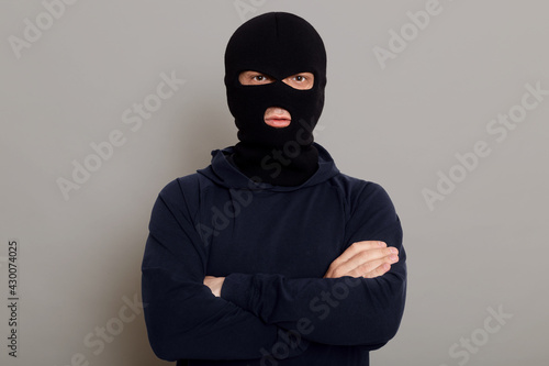 Tableau sur toile Self-confident criminal male posing isolated on a gray background, wearing a black hoodie and a bandit mask, looking at the camera, holding his hands folded on his chest